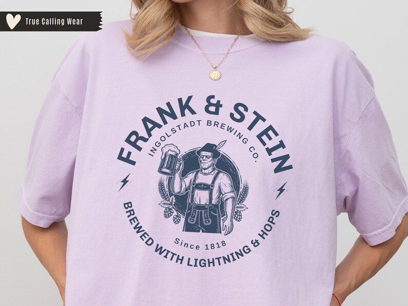 May include: A light purple t-shirt featuring a circular graphic for "Frank & Stein Ingolstadt Brewing Co." The design includes a man holding a beer stein, lightning bolts, and the text "Brewed with Lightning & Hops Since 1818."