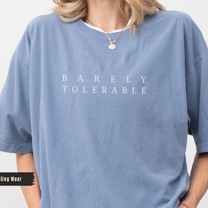 Puede incluir: Camiseta azul claro con las palabras "BARELY TOLERABLE" en letras blancas. La camiseta tiene mangas cortas y cuello redondo. La persona lleva un collar dorado.