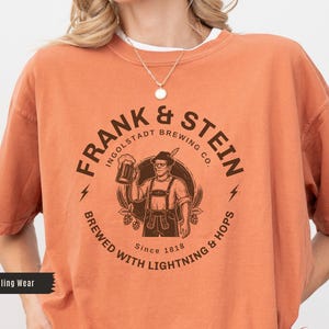 May include: Orange t-shirt with a circular graphic featuring the text "FRANK & STEIN INGOLSTADT BREWING CO." and an illustration of a man holding a beer stein. The shirt also includes the text "BREWED WITH LIGHTNING & HOPS Since 1818."