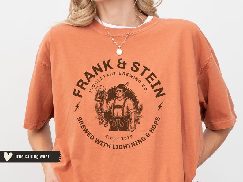 May include: Orange t-shirt with a circular graphic featuring the text "FRANK & STEIN INGOLSTADT BREWING CO." and an illustration of a man holding a beer stein. The shirt also includes the text "BREWED WITH LIGHTNING & HOPS Since 1818."