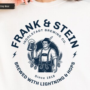 May include: Cream-colored t-shirt featuring a circular graphic with the text "FRANK & STEIN INGOLDSTADT BREWING CO." and a cartoon of Frankenstein holding a beer stein. The design includes lightning bolts, hops, and the text "BREWED WITH LIGHTNING & HOPS Since 1818."