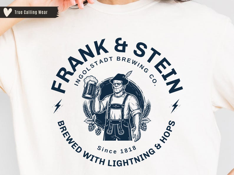 May include: Cream-colored t-shirt featuring a circular graphic with the text "FRANK & STEIN INGOLDSTADT BREWING CO." and a cartoon of Frankenstein holding a beer stein. The design includes lightning bolts, hops, and the text "BREWED WITH LIGHTNING & HOPS Since 1818."