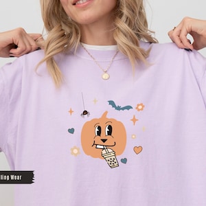 Op de afbeelding: Lichtpaars T-shirt met een cartoon pompoen ontwerp die een drankje drinkt. Het ontwerp bevat een spin, vleermuis, bloemen en harten. De tekst "True Calling Wear" staat onderaan.