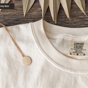 May include: Cream-colored Comfort Colors t-shirt with a gold chain necklace featuring a round pendant. The shirt has a tag that reads "Comfort Colors." The image also includes the text "True Calling Wear."
