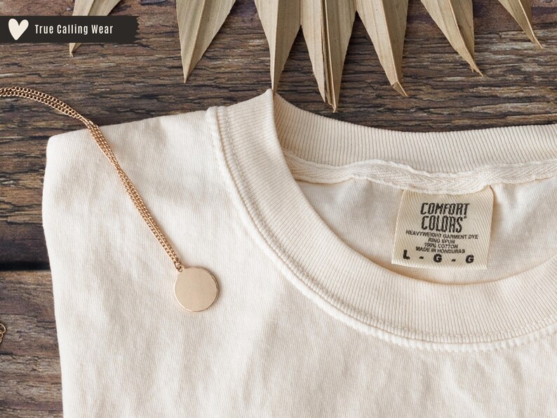 May include: Cream-colored Comfort Colors t-shirt with a gold chain necklace featuring a round pendant. The shirt has a tag that reads "Comfort Colors." The image also includes the text "True Calling Wear."