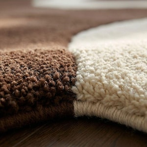 May include: Close-up of a brown and cream-colored rug. The rug has a textured, plush surface with a distinct color block design. The brown section is a darker shade, while the cream section is lighter. The rug appears to be made of soft material.