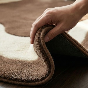 May include: A brown and cream-colored rug with a wavy pattern. The rug is made of a soft, textured material. The edge of the rug is being lifted to show the backing.