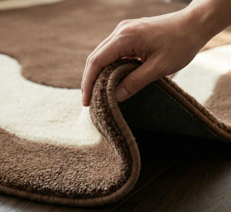 May include: A brown and cream-colored rug with a wavy pattern. The rug is made of a soft, textured material. The edge of the rug is being lifted to show the backing.