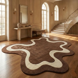 May include: A brown area rug with an abstract shape and a cream-colored wavy design. The rug is placed on a hardwood floor in a room with natural light and a staircase.