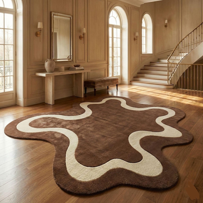 May include: A brown area rug with an abstract shape and a cream-colored wavy design. The rug is placed on a hardwood floor in a room with natural light and a staircase.