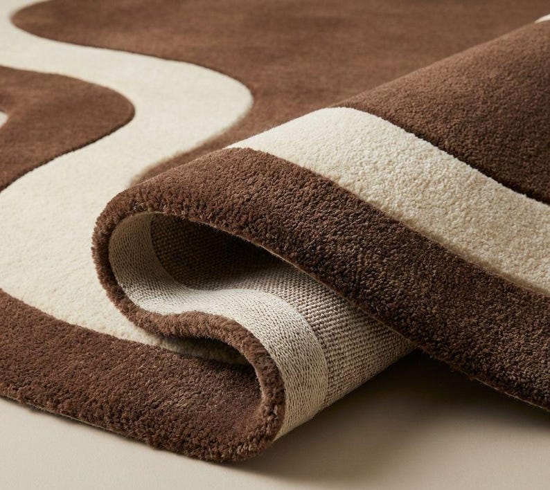 May include: A rolled-up brown and cream-colored rug. The rug features a wavy cream design against a brown background. The edges are also brown, creating a border. The rug is made of a soft, plush material.