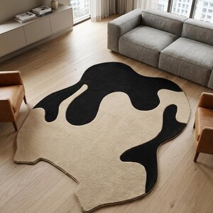 Könnte beinhalten: Ein moderner Teppich mit einem abstrakten Design in Schwarz und Beige. Der Teppich hat eine einzigartige, fließende Form. Er liegt auf einem Holzboden und ist von Möbeln umgeben, darunter ein graues Sofa und zwei braune Sessel.