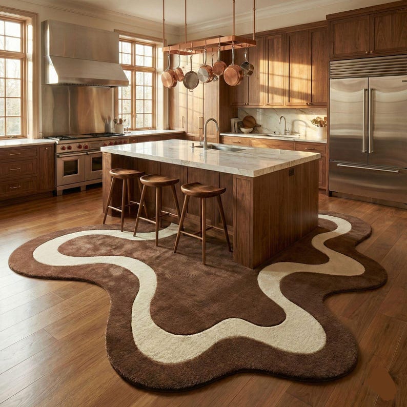 May include: A modern kitchen with a large island, wooden stools, and a unique brown and cream-colored rug. Copper pots hang above the island. The kitchen features stainless steel appliances and wooden cabinetry.