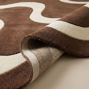 May include: A rolled-up brown and cream-colored area rug. The rug features a wavy pattern in cream against a brown background. The underside of the rug is visible, showing a woven texture. The rug's edges are neatly finished.