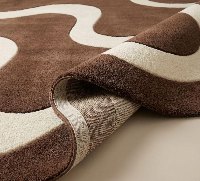 May include: A rolled-up brown and cream-colored area rug. The rug features a wavy pattern in cream against a brown background. The underside of the rug is visible, showing a woven texture. The rug's edges are neatly finished.