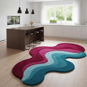 Könnte beinhalten: Ein moderner, abstrakter Teppich mit wellenförmigem Design in den Farbtönen Burgunderrot, Rosa, Hellblau und Türkis. Der Teppich liegt auf einem hellen Holzboden in einer modernen Küche. Die einzigartige Form des Teppichs sorgt für Farbe und visuelles Interesse.