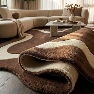 May include: A rolled-up brown and cream-colored area rug with a wavy design. The rug is made of a soft, plush material and is placed on a wooden floor. A beige sectional sofa and marble coffee table are in the background.