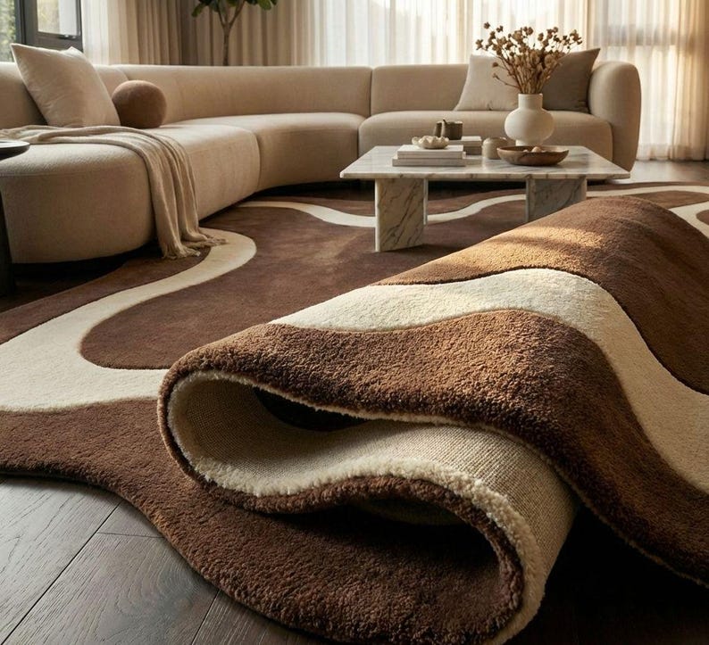 May include: A rolled-up brown and cream-colored area rug with a wavy design. The rug is made of a soft, plush material and is placed on a wooden floor. A beige sectional sofa and marble coffee table are in the background.