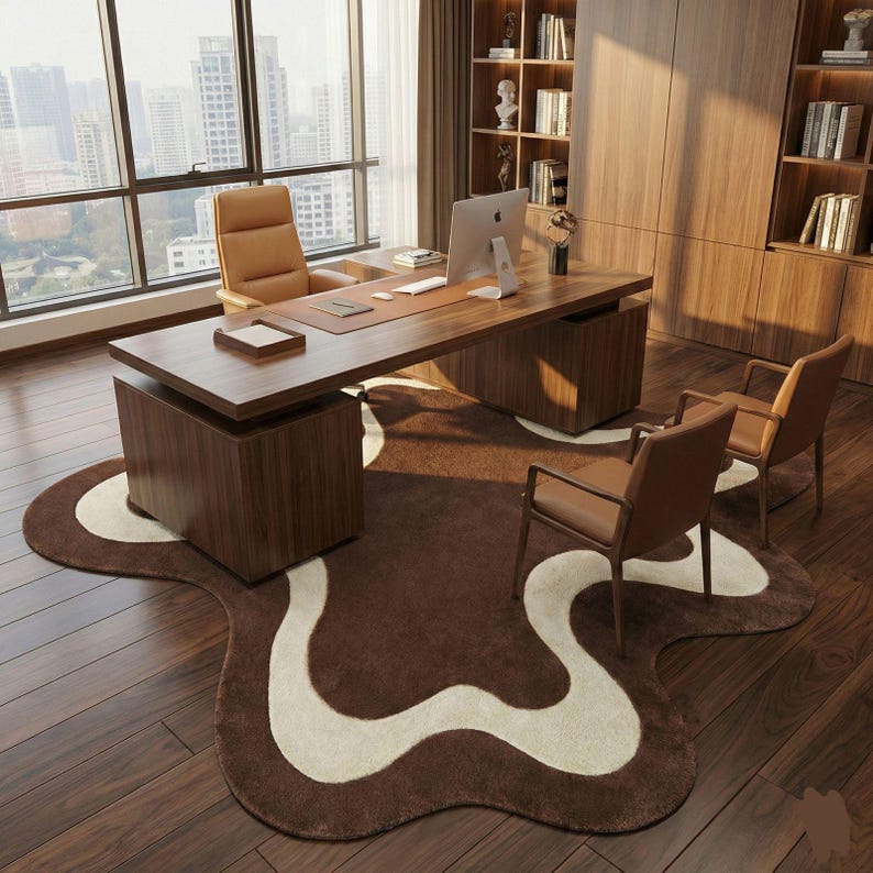 May include: A modern office setup featuring a large wooden desk with a leather top, a matching chair, and two guest chairs. A brown and white patterned rug sits on a hardwood floor. A computer monitor and bookshelves are visible.