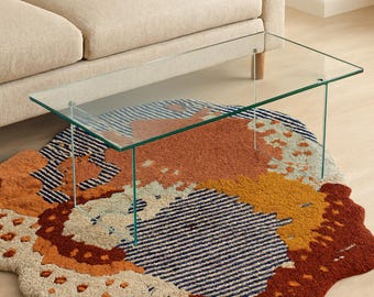 May include: A modern living room setup featuring a beige sofa, a clear glass coffee table, and a colorful rug. The rug has an abstract design with shades of orange, brown, blue, and beige. The coffee table is rectangular and supported by thin, clear legs.