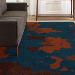 May include: A rectangular rug featuring a dark teal base with abstract rust-coloured shapes. The rug is placed on a wooden floor, with a grey sofa and a wooden console table in the background. The rug's design adds a modern touch to the room.