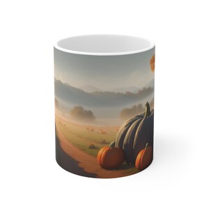 May include: A white ceramic mug with a scenic autumn scene printed on it. The scene features a foggy field with a dirt road leading to a line of trees in the distance. There are several pumpkins scattered throughout the field, including three large pumpkins in the foreground.