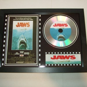 May include: A framed silver disc featuring a Jaws movie poster and a Jaws logo. The disc has a silver background with a blue and white image of a shark's mouth. The poster displays the Jaws movie title and the names of the actors and director. The Jaws logo is in a black and white film strip frame.