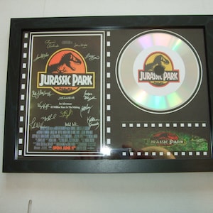 Puede incluir: Un póster enmarcado en blanco y negro de la película Jurassic Park con un disco plateado que presenta el logotipo de Jurassic Park. El póster tiene el título de la película, la fecha de estreno y una lista de nombres de actores. El disco está enmarcado en un marco negro con un borde de tira de película.