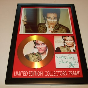 Puede incluir: Un disco de oro en un marco rojo con un paspartú negro. El disco presenta una foto de Adam Ant. El marco también incluye una foto de la portada del álbum de Adam Ant y un autógrafo firmado. El texto "LIMITED EDITION COLLECTORS FRAME" está impreso en la parte inferior del marco.