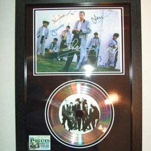 May include: A framed black and white photo of a band with their signatures. The band is standing in front of a white wall. The photo is accompanied by a silver platinum record with the band's name and album title, "The Pogues Dirty Old Town", printed on the label.