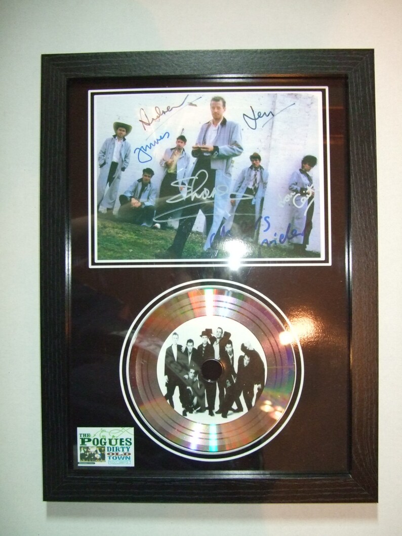 K&ouml;nnte beinhalten: Ein gerahmtes Schwarzwei&szlig;foto einer Band mit ihren Unterschriften. Die Band steht vor einer wei&szlig;en Wand. Das Foto wird von einer silbernen Platin-Schallplatte mit dem Namen der Band und dem Albumtitel "The Pogues Dirty Old Town" begleitet, der auf dem Etikett gedruckt ist.