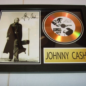 May include: A framed gold CD featuring a black and white image of Johnny Cash playing a guitar. The CD is set within a black frame with a gold plaque that reads "JOHNNY CASH".