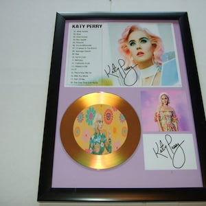 May include: A gold record award with a floral design and a picture of Katy Perry. The award is framed with a black frame and a purple mat. The frame also includes a picture of Katy Perry and her signature.