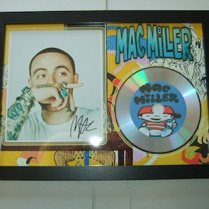 Op de afbeelding: Een ingelijste zilveren cd met een kleurrijk cartoonfiguur op de voorkant. De cd is getiteld "Mac Miller". De lijst bevat ook een zwart-witfoto van Mac Miller met zijn handtekening.