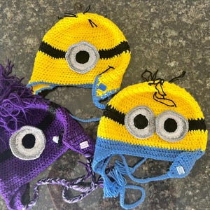 May include: Three handmade crochet hats in the style of Minions. Two are yellow with black stripes and blue trim, and one is purple with black stripes. Each hat features a large eye or two eyes, and some have dangling yarn.