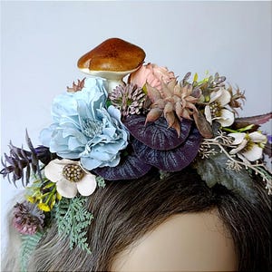 May include: A floral crown with a brown mushroom, light blue and pink flowers, and various greenery. The crown includes purple leaves and small white flowers, creating a fairy-tale aesthetic. Ideal for themed events or costume parties.