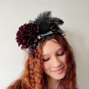 Könnte beinhalten: Ein dunkelburgunderfarbener floraler Haarschmuck mit einer großen, geschichteten Blume und schwarzen Federn. Der Haarschmuck hat auch kleinere Blumen und Blätter in Grau- und Burgundertönen. Das Design ist vor einem weißen Hintergrund.