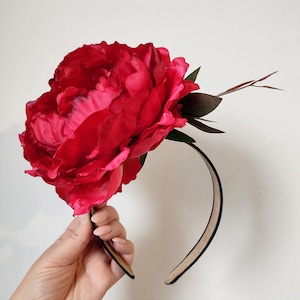 Op de afbeelding: Een felrode pioenroos bloemen haarband. De grote, gekreukelde bloem is het middelpunt, aangevuld met donkergroene bladeren en slanke bruine stelen. De haarband zelf is zwart met een bruin interieur, wat een opvallend contrast creëert.