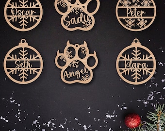 May include: Six wooden Christmas ornaments with snowflake designs.  Two ornaments are round with the names "Oscar", "Seth", "Peter", and "Klara" written on them. Two ornaments are paw-shaped with the names "Sadie" and "Angel" written on them.