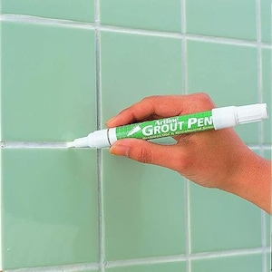 Op de afbeelding: Een witte voegenpen met een groen label waarop "Artline Grout Pen" en "Restores Old & Discoloured Grout" staat. De pen wordt door een hand tegen een betegelde muur gehouden.
