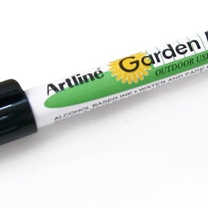 Peut inclure: Marqueur de jardin noir et vert avec le texte "Artline Garden Marker" et "Outdoor Use Xylene Free" et "Alcohol Based Ink Water and Fade Resistant".