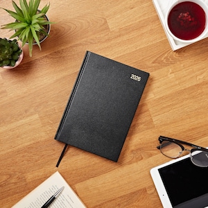 Könnte beinhalten: Ein schwarzer 2026er Tagesplaner mit einem goldenen, geprägten Jahr auf dem vorderen Umschlag. Der Planer liegt auf einer Holzoberfläche mit einem Buch, einer Brille, einem Tablet und einer Tasse Tee.