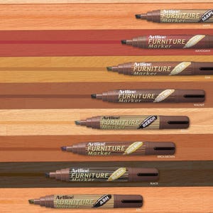 May include: A set of eight wood stain markers, labelled with different wood types: Maple, Mahogany, Oak, Walnut, Beech, Birch Brown, Black, and Ash. The markers are arranged in a row on a wooden surface, with the stain colour visible on the wood.
