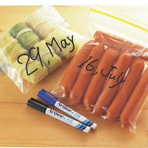 Pode incluir: Duas sacolas de plástico transparentes com comida dentro. Uma sacola contém rolinhos de repolho e está etiquetada com "29. May". A outra sacola contém salsichas e está etiquetada com "16. July". Dois marcadores permanentes pretos e azuis estão sobre uma superfície de madeira.