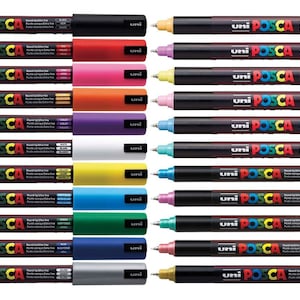 Op de afbeelding: Een verzameling Uni Posca verfmarkers in diverse kleuren, waaronder zwart, rood, roze, oranje, paars, wit, geel, blauw, groen, zilver en goud. Elke marker heeft een zwarte behuizing en een gekleurde punt.