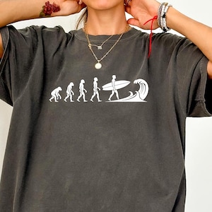 May include: Dark gray t-shirt featuring a white graphic of human evolution ending with a surfer and a wave. The word "PEPPER" is printed in white on the bottom right corner of the shirt. The shirt has short sleeves and a relaxed fit.