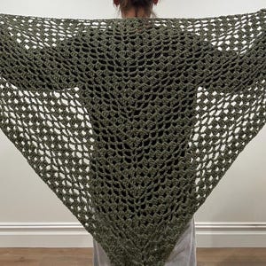 May include: Olive green crochet shawl with a triangular shape. The shawl features a delicate openwork design, creating a lacy texture. The shawl is draped over the shoulders, showcasing its full width and intricate pattern. The color is a muted, earthy tone.