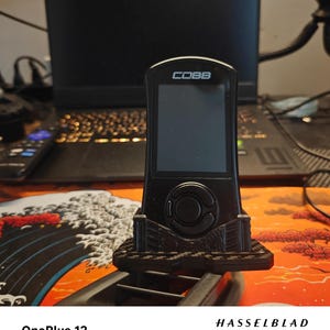 May include: Black electronic device with the word "COBB" on the top. The device has a screen and a circular button. It is mounted on a black stand. The background includes a laptop and a colorful desk mat.