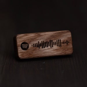 Puede incluir: Una etiqueta rectangular de madera con un logotipo de Spotify y un código de barras en blanco y negro grabado en ella.