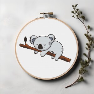 Op de afbeelding: Een witte koalabeer met gesloten ogen slaapt op een bruine tak. De koala bevindt zich in een kruissteek-borduurring.